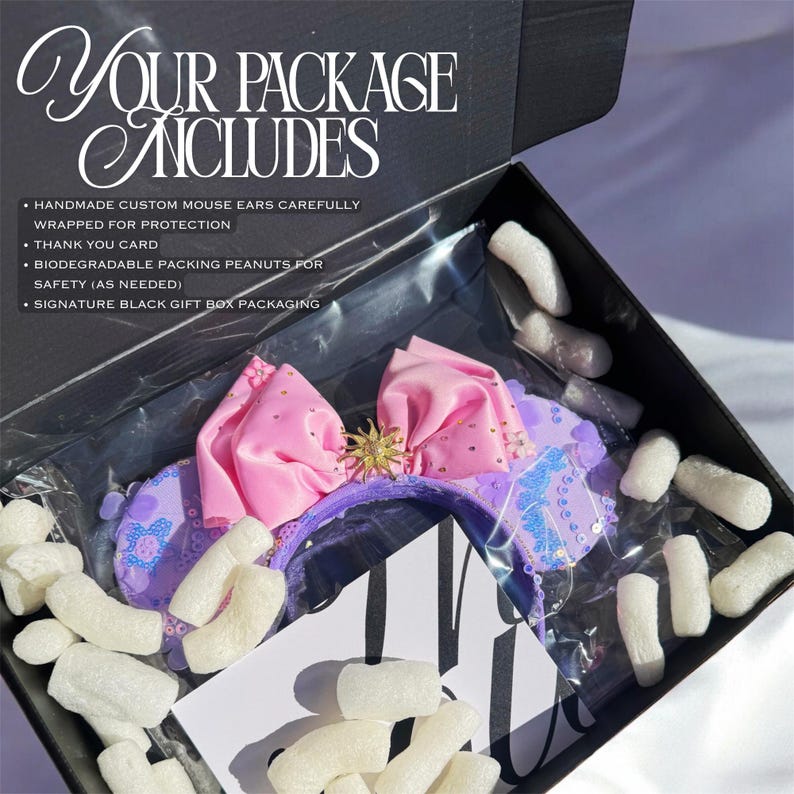 May include: A black gift box containing purple mouse ears with a large pink bow and a gold sun-shaped center emblem. The box includes white biodegradable packing peanuts, a thank you card, and text reading: YOUR PACKAGE INCLUDES, HANDMADE CUSTOM MOUSE EARS CAREFULLY WRAPPED FOR PROTECTION, THANK YOU CARD, BIODEGRADABLE PACKING PEANUTS FOR SAFETY (AS NEEDED), SIGNATURE BLACK GIFT BOX PACKAGING.