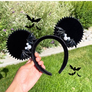 May include: Black velvet mouse ears with bat wing embellishments and pearl accents. The ears are attached to a black headband.