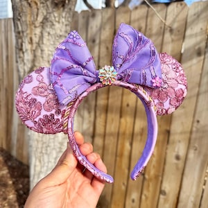 May include: A purple and pink Minnie Mouse ear headband. The ears are a light pink with a floral design. The bow is a darker purple with sequins. A jeweled flower is in the center of the bow. The headband is held by a hand.