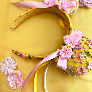 May include: A yellow headband featuring two circular ears decorated with pink and orange floral embroidery, a gold mesh overlay, and light pink satin bows. The ears are accented with small clear rhinestones and additional pink fabric flowers.