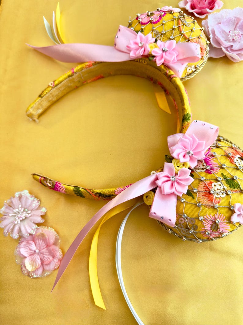 May include: A yellow headband featuring two circular ears decorated with pink and orange floral embroidery, a gold mesh overlay, and light pink satin bows. The ears are accented with small clear rhinestones and additional pink fabric flowers.