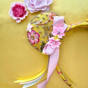 May include: A yellow headband featuring a circular ear covered in gold netting, clear rhinestones, and pink floral embroidery. The ear is adorned with two pink fabric flowers with rhinestone centers and a long, light pink satin ribbon tail.