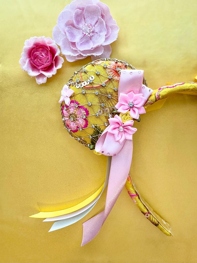 May include: A yellow headband featuring a circular ear covered in gold netting, clear rhinestones, and pink floral embroidery. The ear is adorned with two pink fabric flowers with rhinestone centers and a long, light pink satin ribbon tail.
