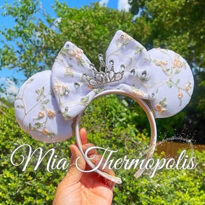 May include: A white headband featuring two round ears and a central bow, all decorated with embroidered floral vines in shades of peach and green. A silver-toned crown ornament with clear crystals sits atop the bow. The text Mia Thermopolis is visible.