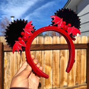 May include: Red headband with black circular accents and red bat wings. The headband features red berries and is designed with a Halloween or gothic theme. The accessory is held up against a wooden fence and a blue sky.