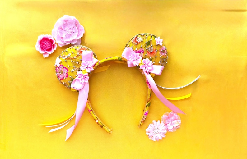 May include: A yellow headband featuring two circular ears covered in yellow fabric with a floral pattern, intricate gold embroidery, and sequins. The ears are decorated with pink fabric flowers, light pink satin ribbons, and dangling yellow ribbons.