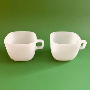 Peut inclure: Deux tasses carrées en céramique blanche avec des anses. Les tasses sont sur un fond vert.