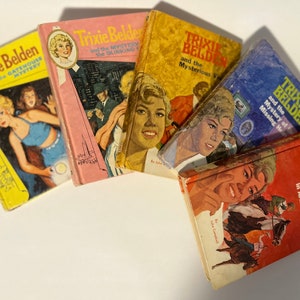 Pode incluir: Uma pilha de cinco livros de bolso com capas coloridas que apresentam uma jovem detetive chamada Trixie Belden. Os livros são intitulados "Trixie Belden e o mistério da casa da porta", "Trixie Belden e o mistério da luz cegante", "Trixie Belden e o visitante misterioso", "Trixie Belden e o mistério do herdeiro desaparecido", e "Trixie Belden e o mistério no Arizona".