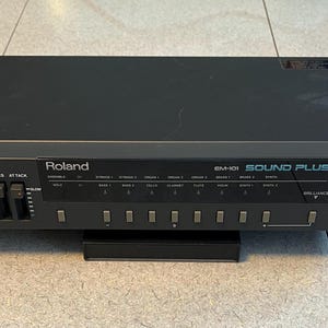 May include: Black Roland EM-101 Sound Plus synthesizer. The front panel features volume, dynamics, and attack controls, along with various sound selection buttons. The unit has a power button and a MIDI message indicator.