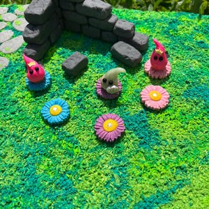 May include: A miniature fairy garden scene with a stone wall, green grass, and three small figurines. Two of the figurines are pink with red crowns and the third is white with black eyes. Each figurine is sitting on a flower with a pink, blue, or purple center.