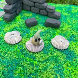 May include: A small, green and white figurine with black eyes, wearing a shell, sitting on a green and blue surface. The figurine is surrounded by three seashells.