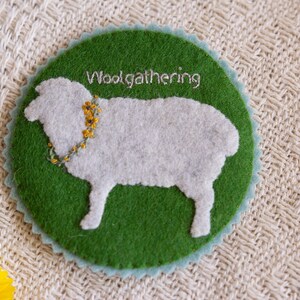 May include: A green felt circle with a white felt sheep silhouette and the text "Woolgathering" embroidered in white thread. The sheep has a yellow flower garland around its neck.