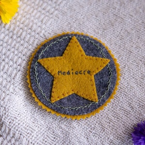 以下が含まれることがあります： 緑色のステッチで囲まれた灰色のフェルトの円の中に、"Mediocre"と刺繍された黄色のフェルトの星がある。