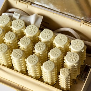 以下が含まれることがあります： 花柄のトップが付いた、クリーム色のヴィンテージ電気ヘアカーラーセット。カーラーは、蓋に鏡が付いたプラスチックケースに入っています。