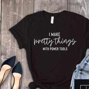 May include: Black t-shirt with white text that reads "I make pretty things with power tools". The shirt is laid flat on a wooden surface with a pair of jeans and shoes beside it. A logo for "Jane Threads everyday tees" is in the bottom right corner.