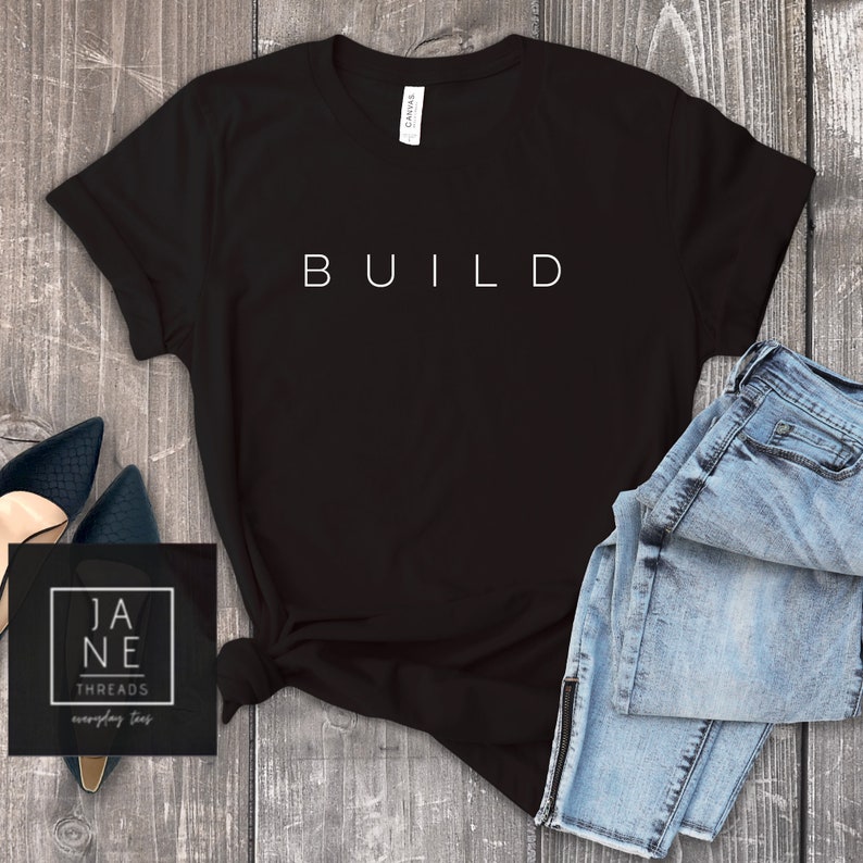 May include: Black t-shirt with the word "BUILD" printed in white on the front. The shirt is laying flat on a wooden surface with a pair of blue jeans and a pair of black shoes.