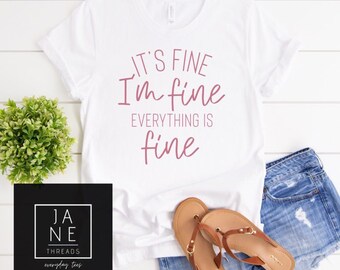 May include: White t-shirt with the text "It's fine I'm fine everything is fine" in pink lettering.