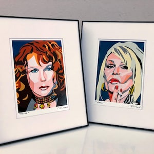 May include: Two framed portrait prints. One features a woman with red hair, blue eyes, and a black choker, labeled "Edina." The other depicts a woman with blonde hair, red lipstick, and a finger to her lips, labeled "Patsy."