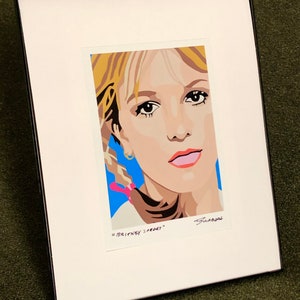 May include: A framed print of a pop art style portrait of Britney Spears. The print is on white paper with a blue background and the text "Britney Spears" is written below the image.