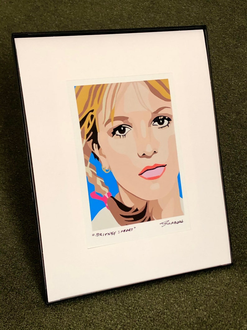 May include: A framed print of a pop art style portrait of Britney Spears. The print is on white paper with a blue background and the text "Britney Spears" is written below the image.