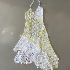 May include: A white and yellow crochet halter dress. The dress features a floral pattern with a scalloped hemline. The halter strap is made of a delicate, crocheted chain.