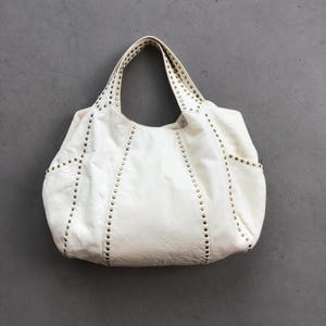 May include: Off-white leather tote bag with studded accents. The bag features two handles and exterior pockets. The studs are evenly spaced along the seams and around the handles. The bag is sitting on a gray surface.