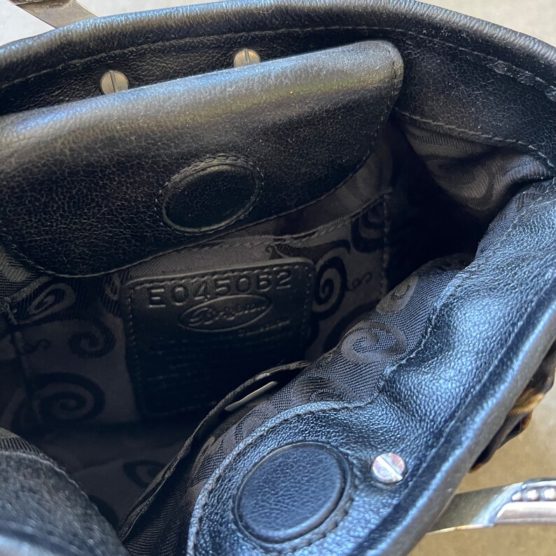 K&ouml;nnte beinhalten: Das Innere einer schwarzen Ledertasche mit schwarzem Stofffutter. Das Futter hat ein Wirbelmuster und der Text "E045062 Fossil" ist auf einem Etikett aufgedruckt.