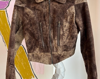 May include: A brown leather jacket with a textured finish, featuring a collar, a front zipper, and two zippered pockets. The jacket is hanging on a metal hanger. The jacket's color is a mix of brown and tan.