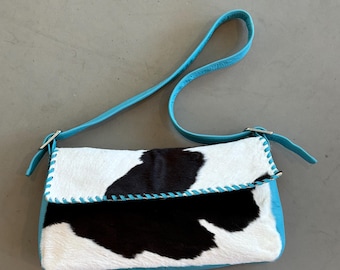 Puede incluir: Bolso rectangular con estampado de piel de vaca en blanco y negro, con correa y ribete de cuero azul turquesa. La solapa está cosida con hilo turquesa y el bolso tiene hebillas plateadas.