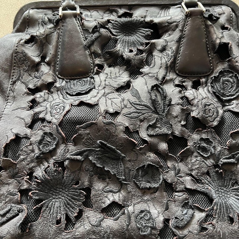 May include: A black leather handbag with a floral cut-out design. The bag has two top handles and a mesh lining.