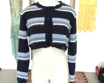 Op de afbeelding: Een cropped vest met horizontale strepen in marineblauw, lichtblauw en wit. De trui heeft lange mouwen en een knoopsluiting aan de voorkant. Het kledingstuk wordt op een mannequin getoond.