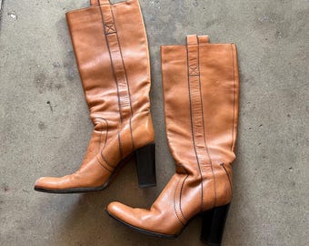 May include: A pair of brown leather knee-high boots with a block heel. The boots have a pointed toe and a decorative stitching detail.