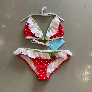 Puede incluir: Conjunto de bikini de dos piezas con estampado de lunares rojos y blancos. La parte superior es de estilo triangular con volantes blancos y detalles verdes. La parte inferior tiene volantes a juego y detalles de cerezas. El bikini está hecho de un material ligero.
