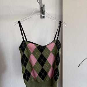 Può includere: Un top a canotta in maglia senza maniche con motivo a rombi nei colori verde oliva, nero e rosa tenue. Il capo presenta sottili spalline nere regolabili, uno scollo dritto e un orlo a coste, il tutto appeso a una gruccia trasparente.