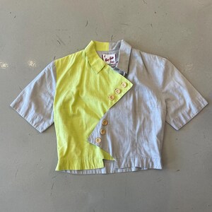 May include: A short-sleeved shirt with a unique design. The shirt is split vertically, with a lime green panel on the left and a gray panel on the right. It features a collar and wooden buttons. The shirt has a tag with text.