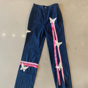 Könnte beinhalten: Dunkelblaue Jeanshose mit hoher Taille und geradem Bein. Die Hose ist mit rosa Bändern und weißen Spitzen-Schmetterlingsapplikationen verziert. Das Design umfasst einen rosa Streifen an der Seite jedes Beins.