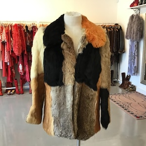May include: A multi-coloured patchwork fur coat on a mannequin. The coat features panels of cream, black, brown, and orange fur. It has a wide collar and long sleeves, with a vintage aesthetic. The coat's design is a striking example of retro fashion.