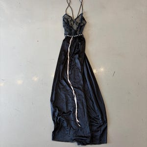 Könnte beinhalten: Schwarzes Satin-Nachthemd mit Spitzenoberteil und Spaghettiträgern. Ein weißer Bandgürtel betont die Taille, und das Nachthemd hat einen langen, fließenden Rock. Das Nachthemd liegt flach auf einer grauen Oberfläche.