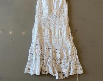 Puede incluir: Un vestido blanco sin tirantes con un diseño de ganchillo. El vestido presenta un corpiño ajustado y una falda escalonada con un dobladillo festoneado. El vestido tiene un cordón en el lateral.