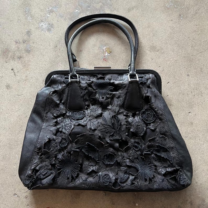 May include: Black leather handbag with a floral design. The bag has a top handle and a clasp closure.