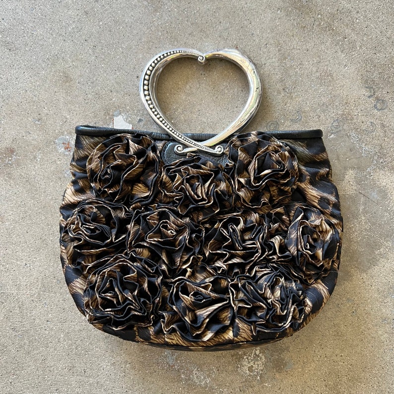 K&ouml;nnte beinhalten: Eine schwarze und braune Handtasche mit einem herzf&ouml;rmigen silbernen Griff und einem Blumendesign.