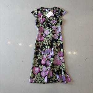 Pode incluir: Um vestido preto floral com decote em V e mangas curtas. O vestido apresenta um padrão de rosas rosa e roxas com folhas verdes. Uma etiqueta está presa na gola. O vestido é feito de um tecido transparente.