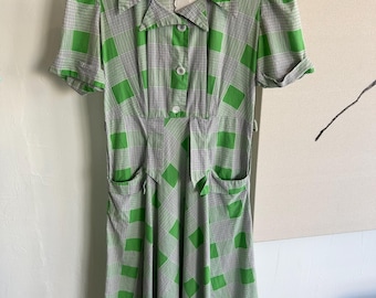 Puede incluir: Un vestido de cuadros verdes y blancos con cuello, mangas cortas y cierre de botones en la parte delantera. El vestido tiene dos bolsillos en la parte delantera y una cintura ajustada.