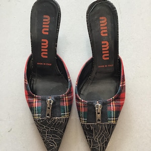 Puede incluir: Un par de mules de tacón Miu Miu. Los zapatos presentan un estampado de cuadros rojos, verdes y amarillos en la parte superior, con una puntera de cuero negro y un detalle de cremallera. La plantilla es negra con el logo de Miu Miu en naranja.