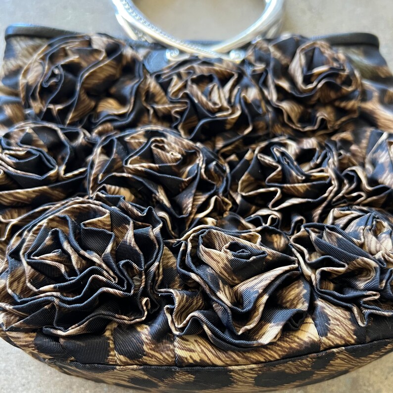 K&ouml;nnte beinhalten: Eine braune und schwarze Handtasche mit einem silbernen Henkel. Die Tasche ist aus Stoff und hat ein Blumenmuster.