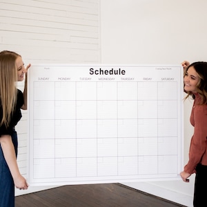 May include: Two people holding up a large white dry-erase calendar with black grid lines and the word "Schedule" printed at the top. The calendar has the days of the week listed across the top: Sunday, Monday, Tuesday, Wednesday, Thursday, Friday, Saturday. The calendar also has the words "Coming Next Month" printed in the top right corner.