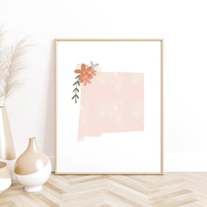 May include: A framed print of the state of New Mexico with a floral design in shades of pink and orange. The print is on a white background and has a light wood frame.