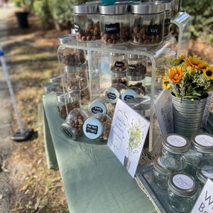 Pode incluir: Um expositor de mesa com pequenos frascos de vidro cheios de sementes marrons, etiquetados com nomes como "Bettoli" e "Kale". Os frascos estão dispostos em uma prateleira de acrílico transparente. Há também frascos de vidro vazios em uma bandeja de metal, e uma placa que diz "Construa seu próprio jardim".