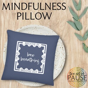 May include: A blue square pillow with white text and a diagram for box breathing. The text reads "Breathe In", "Hold", "Breathe Out", "Hold", and "box breathing". The pillow is on a white plate with a brown wooden background.