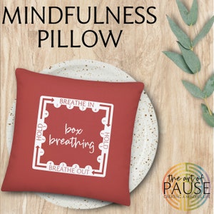 May include: Red square pillow with white text and a diagram for box breathing. The text says "Breathe In", "Hold", "Breathe Out", and "box breathing". The pillow is on a white plate with a wooden background.